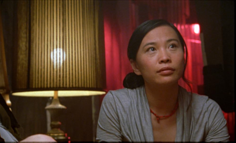 A scene of Sophia Lin in Shortbus (2026) played by Sook-Yin Lee. The Chinese-Canadian actress wears simple grey clothing and looks pensively upwards against a simple domestic background.